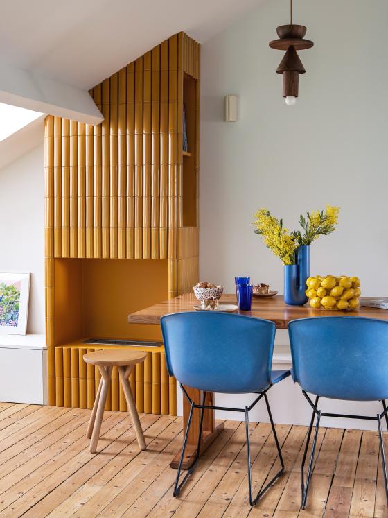 Salon avec cheminée recouverte de faïence, table et chaises colorées.
